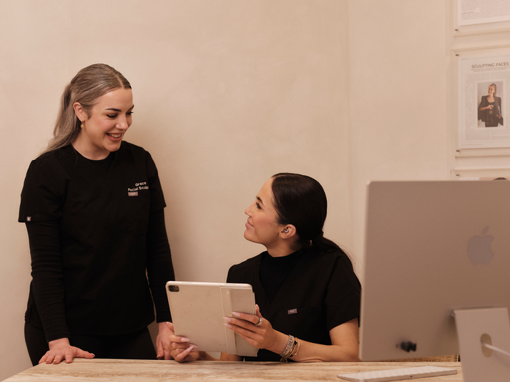 Two practitioners, one sitting down and one standing, smiling at each other. The seated practitioner is holding a tablet.